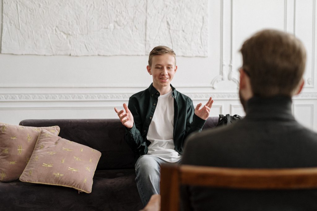 A younger white male sits on a couch talking to an counsellor.