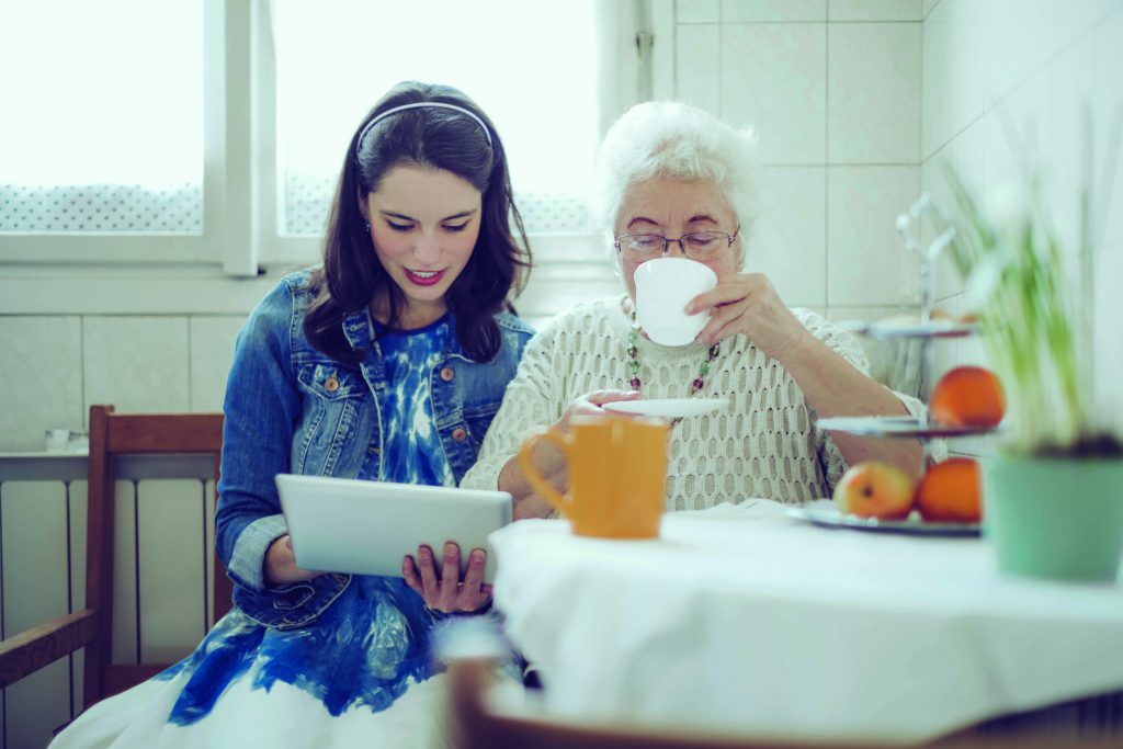 An older white woman drinks from a tea cup while a younger white woman sits at her side showing her something on a tablet.