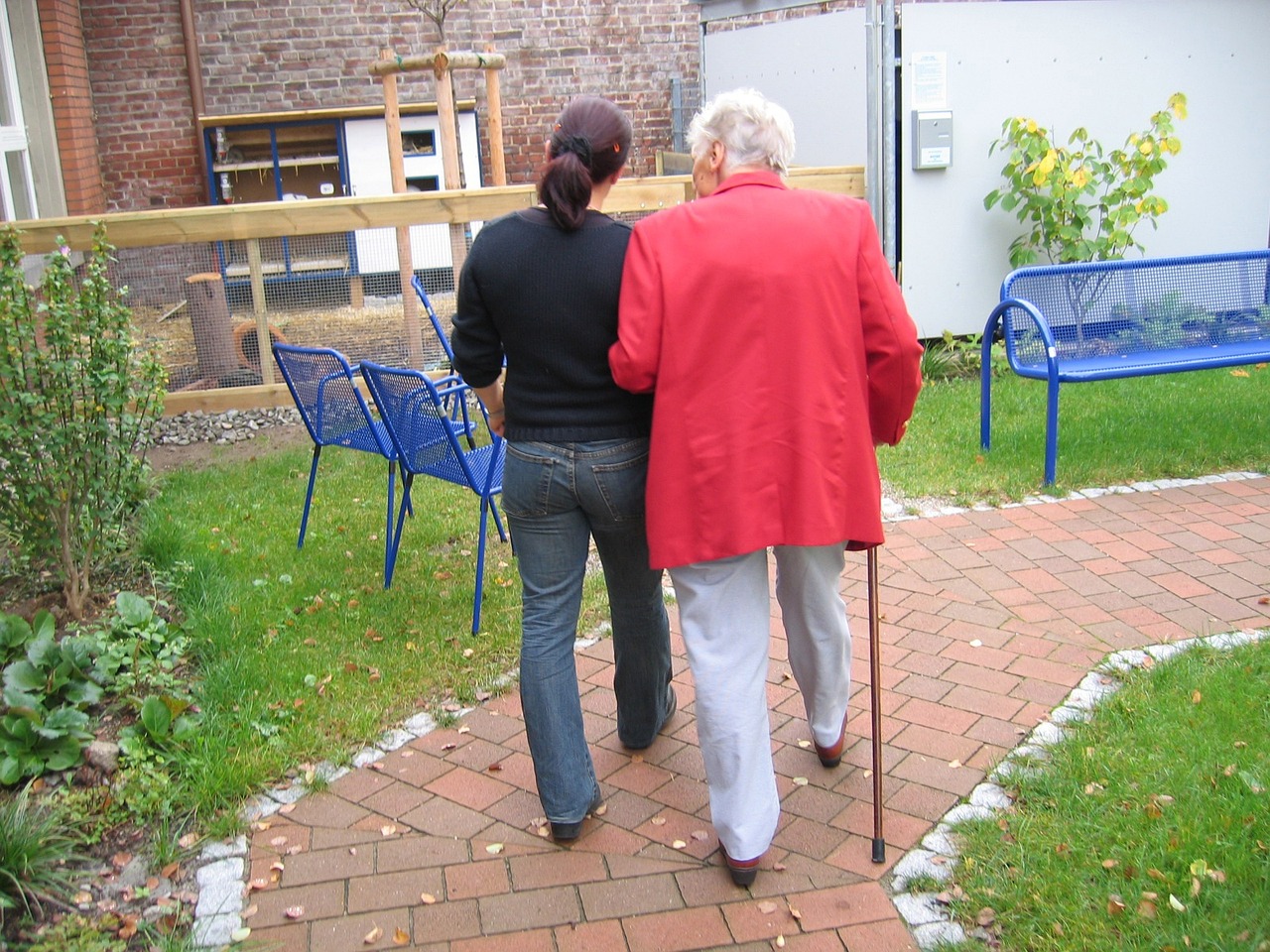 A woman in a red jacket walks with a stick supported by a younger woman. Photo by Geralt on Pixabay,