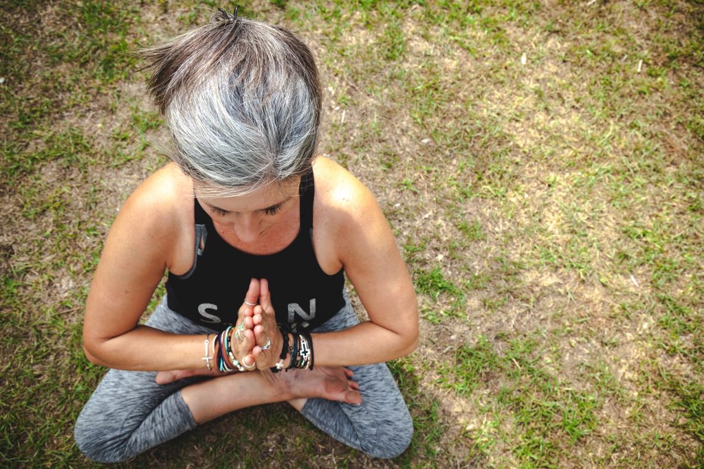 White woman with grey hair sites in a yoga pose on grass. Photo by Kelly Newton on Unsplash
